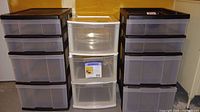 Photo of all three plastic storage drawer units arranged side by side against a wall, showing two black framed units with four drawers each and one white framed unit with three drawers.