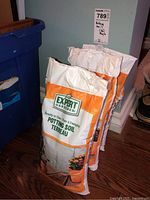 Photo showing three 3L bags of Expert Gardener potting soil standing upright on a wooden floor near a wall corner.