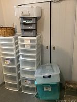 Wide shot showing two tall white plastic storage drawer units with eight drawers each, a gray smaller plastic drawer unit on top, several additional plastic bins stacked, a woven basket, and a small wooden step stool.