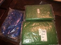 Two green tarps and one blue hose sealed in original plastic packaging, lying on a wooden surface.