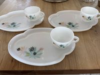 Three white luncheon plates with molded thumb grips and matching cups on top, all with mid-century floral pattern in pastel colors and black, placed on wooden table.