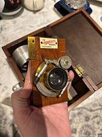 Camera held in hand showing wooden body, brass fittings, lens, and identification plate 'The Sanderson Camera H.T.T. London'.