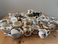Full view of collection of antique and vintage creamers, sugar containers, and small pitchers on wooden table showing floral and gold designs.