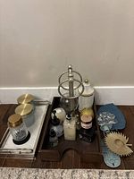 Photo of bathroom decor items arranged on the floor including glass containers with brass lids, wooden and marble trays, lotion and soap dispensers, and decorative items
