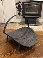 Front quarter view showing the decorative embossed pattern on the log cradle and the ornate handle against background of wood floor and fireplace.