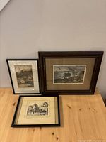 Three framed historical prints leaning against a beige wall on a wooden floor, showing coastal and architectural historical scenes.