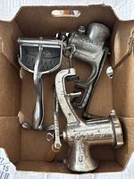 Box containing two metal manual meat grinders and one hand crank juicer, viewed from above