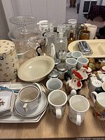 Photo of assorted cups, mugs, ceramic and glass serving dishes, decorative containers, and small novelty salt and pepper shakers arranged on a wood table.