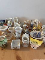 Various miniature ceramic and porcelain collectibles arranged on a table showing pitchers, mugs, small figurines, dishes, and decorative items.