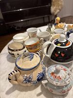 Photo showing close-up of various miniature teapots and teacups with floral, bird, and graphic designs on marble table surface.