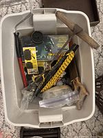Top view of a white plastic bin containing various tools and hardware: hammer, saw, paint roller, rope, Bosch planer box, and plastic bags with assembled hardware items.