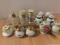 Photo showing full assortment of salt and pepper shakers arranged on wooden surface including clear glass, floral ceramic, and small decorative shakers.