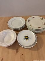 Stack of mixed pattern plates including white plates with embossed floral edges, bee motif plates, and plates with small colorful fruit pattern.