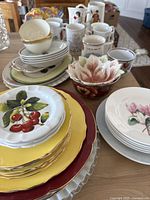 Variety of plates, bowls, mugs, and teacups arranged on table, showing different designs including cherries and flowers