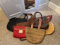Photo shows 7 assorted bags including black leather, brown suede with braiding, woven leather handbag, plaid fabric tote, red nylon pouch, natural woven straw tote, and tan leather bag all displayed on carpet