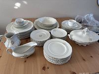 Wide view of the complete antique semi-porcelain English china set arranged on a wooden table, showing stacks of plates, bowls, cups, a gravy boat, creamer, and covered serving dish all in white with gold rims and floral decorations.