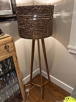 Wooden tripod standing lamp shown fully assembled and lit, placed on a hardwood floor next to furniture.