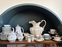 Overall view of antique tea set including large ceramic pitcher, teacups, saucers, and plates arranged on a wooden shelf with blue background