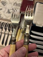 Close-up of two vintage 1940s pastry forks showing bone-colored handles and metal prongs with decorative metal banding, one fork marked with hallmark.