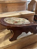 Wooden foot rest showing curved legs and floral tapestry upholstery, with visible wear on edges.
