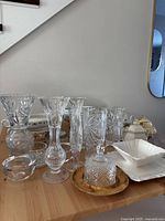Various cut crystal and glass vases and candle holders displayed on a wooden table with some white ceramic dishes in the background.
