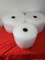 Three large rolls of clear industrial bubble wrap on cardboard tubes positioned on a red fabric surface