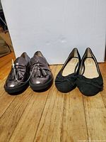 Two pairs of ladies flat shoes, shiny pewter penny loafers with tassels on left and black suede flats with bows on right, placed side by side on wooden floor.