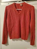 Front view of rust colored V-neck cable knit sweater on hanger showing knit pattern and ribbed cuffs and hem.