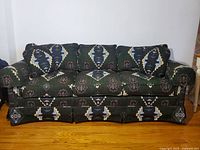 Sofa with dark green geometric patterned fabric and 4 matching throw pillows, positioned against a white wall on a hardwood floor.