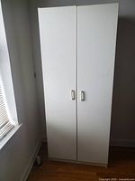 Front view of the tall white wooden storage cabinet with closed double doors and two vertical silver handles.