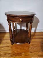 Front view of oval cherry wood end table with pull-out drawer closed, showing round brass knob and turned legs.