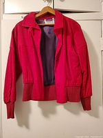 Front view of red corduroy woman's jacket with collar, visible navy blue satin lining inside, long sleeves with ribbed knit cuffs, gold buttons, and knit bottom border.