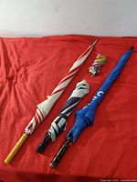 Four umbrellas laid on a red cloth showing different styles including a red and white striped with yellow handle, navy and white striped, rainbow swirl pattern folded compact, and blue umbrella with black handle