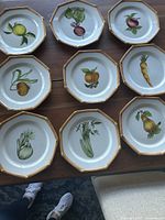Nine octagonal plates with different hand painted fruit and vegetable motifs arranged on a table.