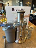 Photo shows Breville Juice Fountain Elite juicer with gray pulp container, clear juice pitcher, and tamper tool in front of the branded box on kitchen counter.