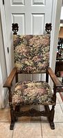 Front view of antique wooden armchair with leaf pattern tapestry upholstery, showing carved details on armrests and frame.