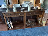 Photo of wood console table placed behind a couch in a living room setting. The console features four small drawers on top and two open storage compartments at the bottom. Various decorative items sit on top of the console but are not included in the lot.
