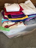 Plastic container filled with various folded women's casual wear including tops in multiple colors such as white, red, purple, and turquoise.