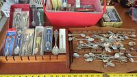 Wide view of two wooden holders filled with boxed and loose collectible souvenir spoons on a table, also showing an adjacent plastic storage bin.