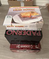 Stacked view of three boxed cookware sets showing Starfrit Heat Magic on top, Paderno cookware set in the middle, and a 7-piece cookware set at the bottom