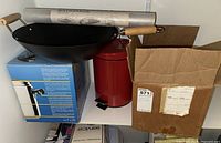 Two boxed pressure cookers, a black wok with wooden handles, roll of shelf liner, and red step-on waste bin on shelf with cardboard box.