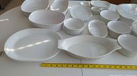 Multiple white casserole dishes and serving plates arranged on a table with a measuring tape for scale.