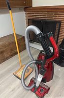 Samsung PowerJet red canister vacuum cleaner with flexible gray hose, black handle, and accompanying carpet sweeper with wooden and yellow handle near a fireplace.