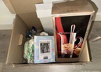 Box containing various home decor items including a boxed pitcher set and a picture frame.