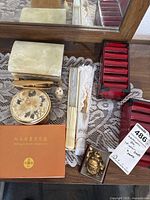 Overhead photo showing a collection of various decorative and functional trinket boxes and items displayed on a lace tablecloth, including the mahogany name card box, floral round box, puff dispenser, fans, small statue, and red container.