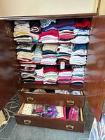 Full view of wooden cabinet with closet door open showing multiple shelves stacked with folded women’s tops and two drawers open holding scarves and sachets.