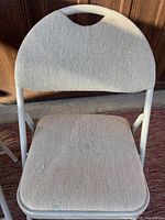 Front and slightly angled top view of one upholstered folding chair showing textured light neutral upholstery and metal frame with carrying handle integrated into backrest.