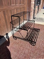 Front perspective photo showing the black metal 3-tier stair step plant stand sitting on a carpeted porch area with a wooden garage door in the background. Shows lattice style tiers and curved feet.
