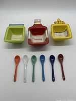 Photo of three colorful square condiment bowls with matching labels and six colorful ceramic spoons laid beneath them.