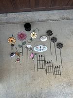 Photo showing various colorful and black metal garden stakes shaped like flowers and birds, hanging decorations, small black garden fence panels, a small black pot, and decorative stones spread on a concrete floor.
